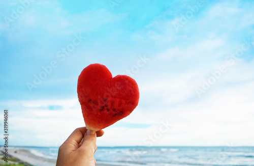 Happy valentines Day or summer holiday creative concept  hand holding heart shaped watermelon against blue sky and beach soft focus background.