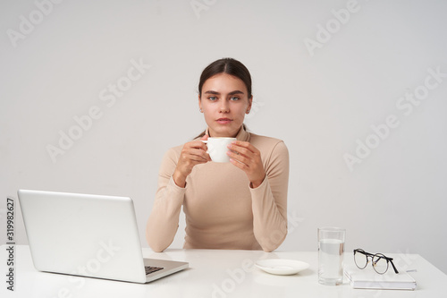 Charming young beautiful blue-eyed brunette lady holding cup of tea in raised hands and looking at camera with calm face, dressed in formal clothes while posing over white background