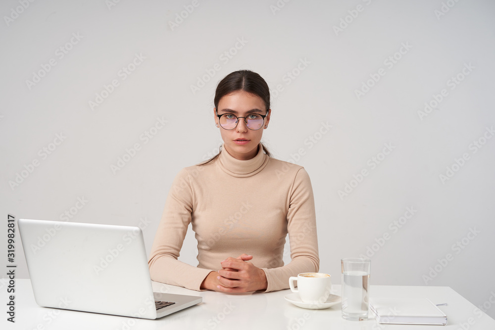 Serious young dark haired businesswoman wearing eyewear and formal clothes while sitting at table with laptop, folding hands on countertop while posing over white background Serious young dark haired businesswoman wearing eyewear and formal clothes while sitting at table with laptop, folding hands on countertop while posing over white background