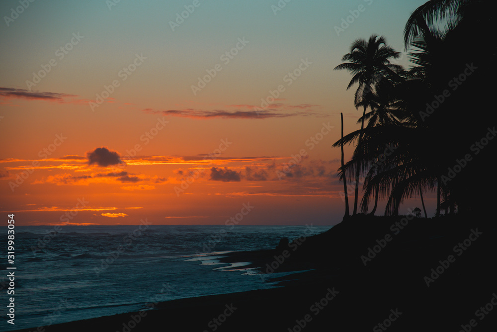 Sun is rising on the beach of a tropical island. Pink and orange sky, palm trees, waves: a postcard from paradise. Scenic view, vacation concept.
