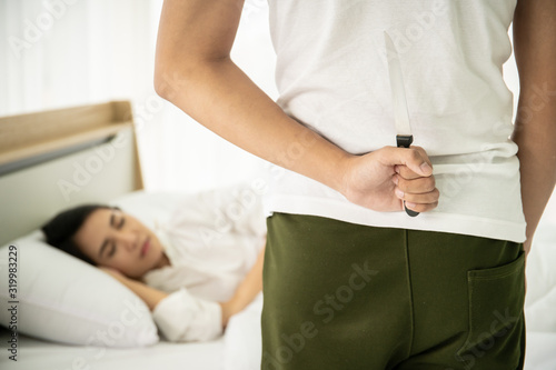 Man hiding a knife behind himself in front of his wife who sleeping on the bed. Concept of unfaithful couple relationship.