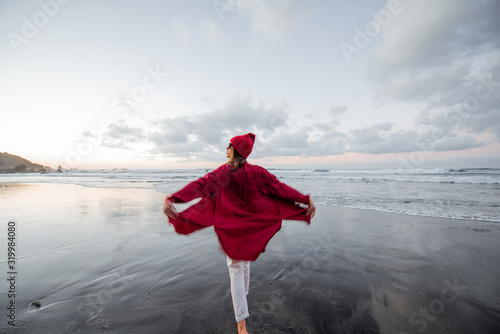 Lifestyle portrait of a carefree woman dressed in red shirt and hat walking on the beach at dusk. Wellness, happiness and life enjoyment concept