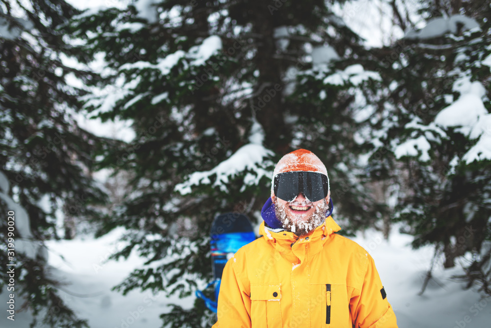 Cheerful smiling snowboarder with snow on his beard and hat stay in yellow jacket in the winter forest on powder day. Cheerful smiling snowboarder with snow on his beard and hat stay in yellow jacket in the winter forest on powder day.