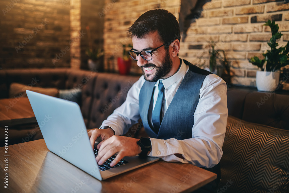 Happy dedicated handsome bearded Caucasian businessman in suit and with eyeglasses sitting in cafe and using laptop for work. Happy dedicated handsome bearded Caucasian businessman in suit and with eyeglasses sitting in cafe and using laptop for work.