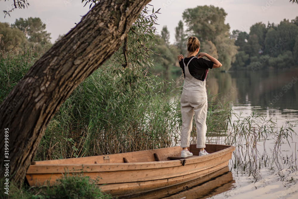 Young woman in a beige overalls stands in wooden fishing boat. Sunset over the river in autumn. Romantic view of boat in the pond and forest.