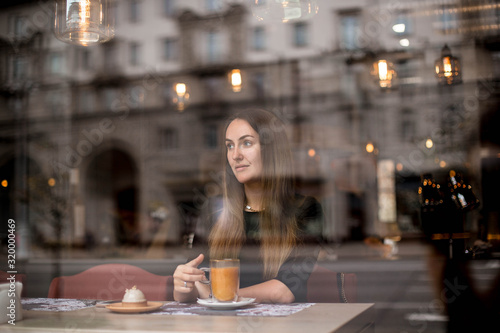 Beautiful girl with long hair drinks orange sea buckthorn tea in cafe behind the window. City street is reflected in the window. Girl in a cafe through the glass.