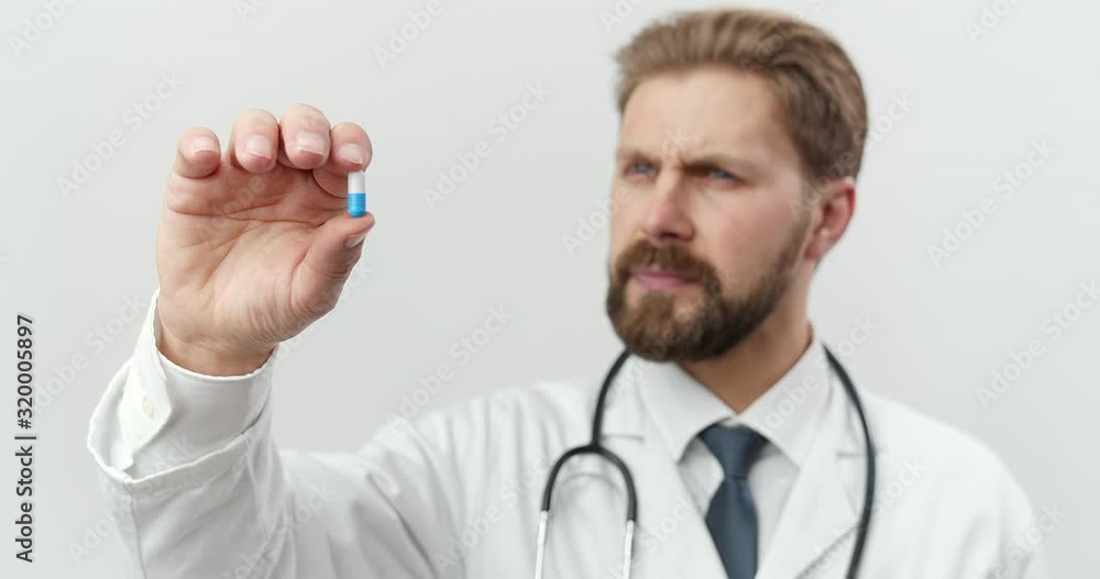 Serious bearded medical expert in white lab coat and black tie with stethoscope on neck examining pill capsule that holding in hand. Isolated over white studio background. Serious bearded medical expert in white lab coat and black tie with stethoscope on neck examining pill capsule that holding in hand. Isolated over white studio background.