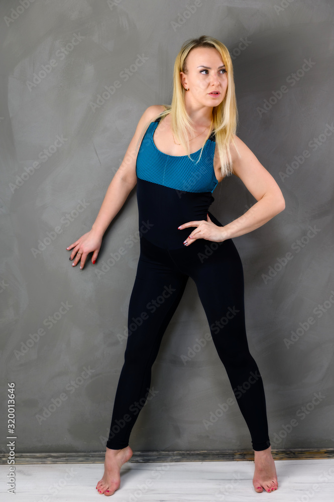 The concept of one fitness. Full-length photo of a pretty slender blonde woman athlete in a tracksuit on a gray background. Standing in different poses with emotions and a smile. The concept of one fitness. Full-length photo of a pretty slender blonde woman athlete in a tracksuit on a gray background. Standing in different poses with emotions and a smile.