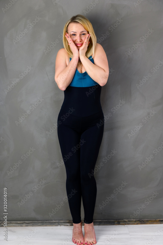 The concept of one fitness. Full-length photo of a pretty slender blonde woman athlete in a tracksuit on a gray background. Standing in different poses with emotions and a smile. The concept of one fitness. Full-length photo of a pretty slender blonde woman athlete in a tracksuit on a gray background. Standing in different poses with emotions and a smile.