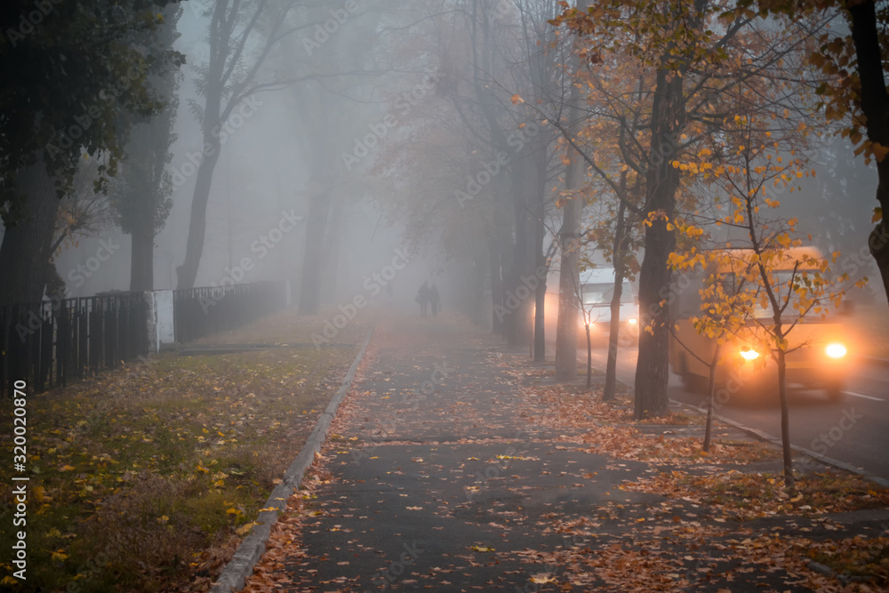 Cars move along an autumn road in conditions of poor visibility. Blurred fog effect. The sidewalk with a pair of walkers is in dense fog. Bad weather on city street.