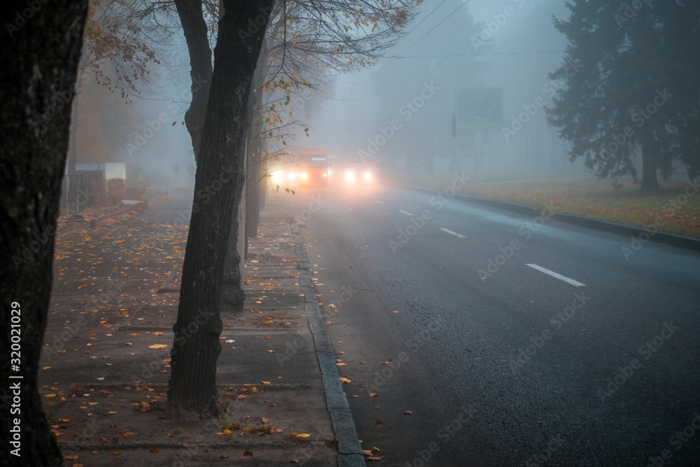 Obraz premium Heavy fog on the road in the early morning. City street with cars in the distance is in poor visibility. The bright light of the headlights of the car. Blurry fog effect.