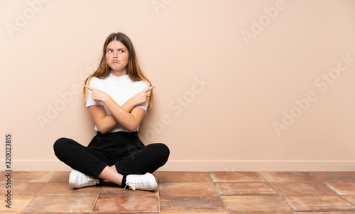 Ukrainian teenager girl sitting on the floor pointing to the laterals having doubts