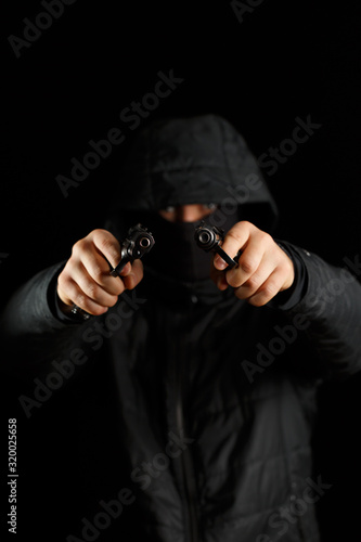 A man with two pistols on a black background. The gun is in focus, the masked man is blurred.
