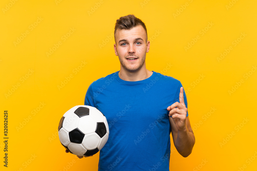 Young handsome blonde man holding a soccer ball over isolated yellow background intending to realizes the solution while lifting a finger up