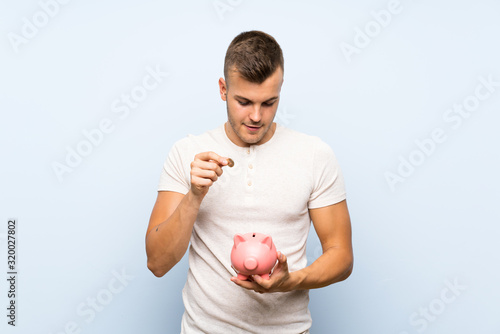 Young handsome blonde man over isolated blue background holding a big piggybank