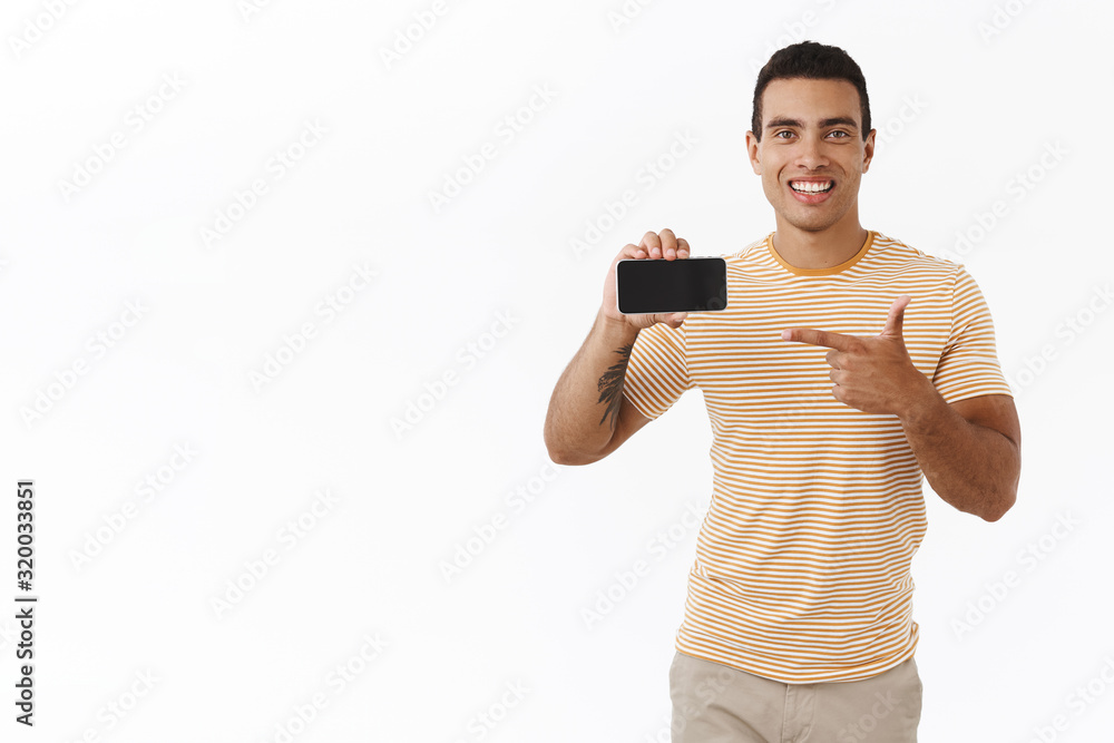 Happy, outgoing handsome athletic man in t-shirt, holding smartphone horizontally and pointing finger at display, recommend good mobile game, useful application, white background Happy, outgoing handsome athletic man in t-shirt, holding smartphone horizontally and pointing finger at display, recommend good mobile game, useful application, white background