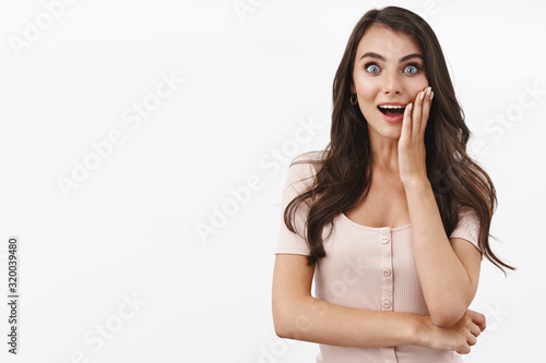 Surprised and impressed, wondered pretty brunette caucasian female, hearing amazing news, open mouth fascinated, touch cheek from amusement, looking curious camera, smiling, white background