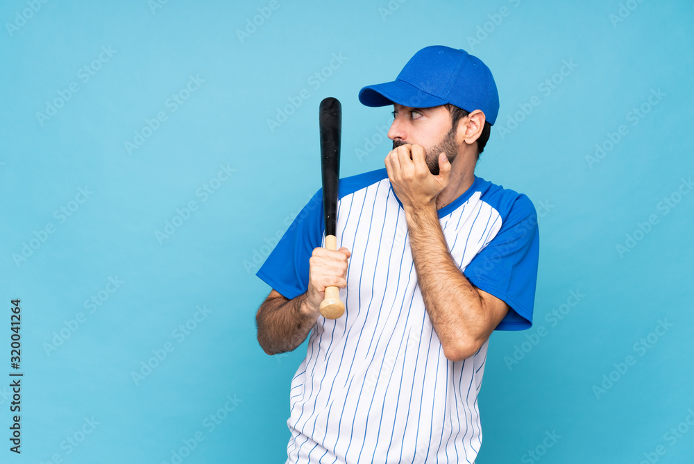 Young man playing baseball over isolated blue background nervous and scared putting hands to mouth