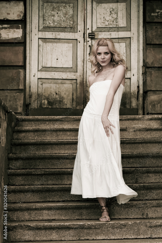 Black-and-white photo of a beautiful young woman in a white dress descends the stairs on the background of an old building with doors. Gothic romance concept Black-and-white photo of a beautiful young woman in a white dress descends the stairs on the background of an old building with doors. Gothic romance concept