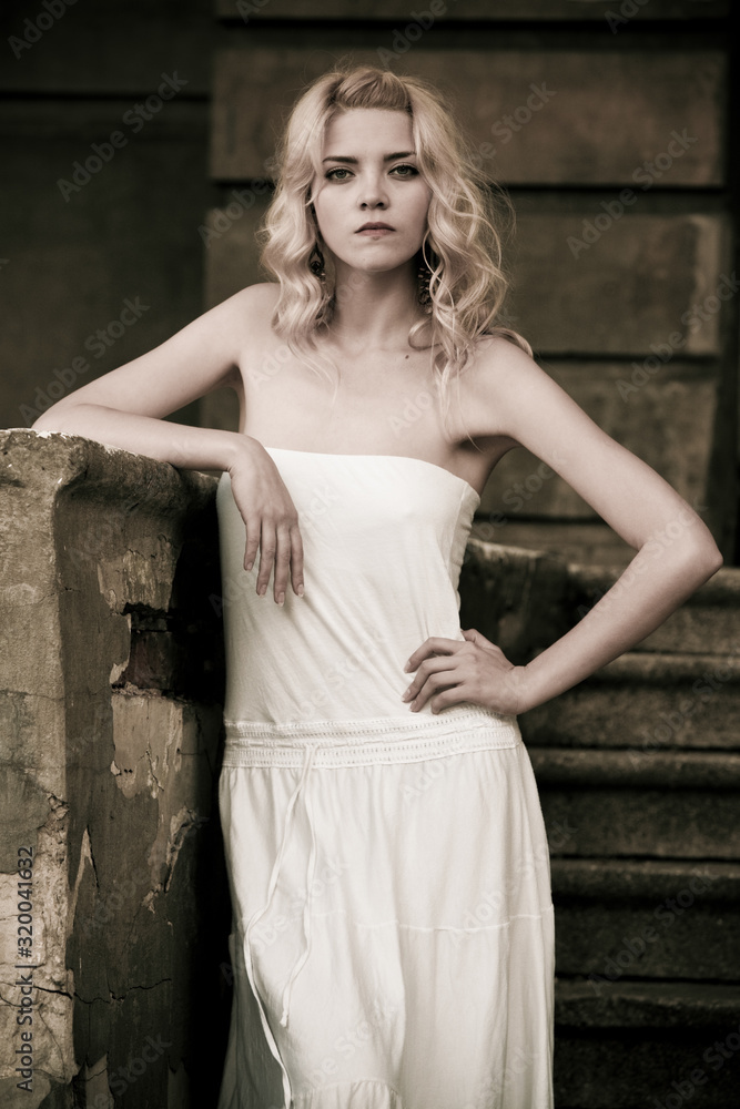 Naklejka premium Black-and-white photo of a beautiful young woman in a white dress descends the stairs on the background of an old building with doors. Gothic romance concept