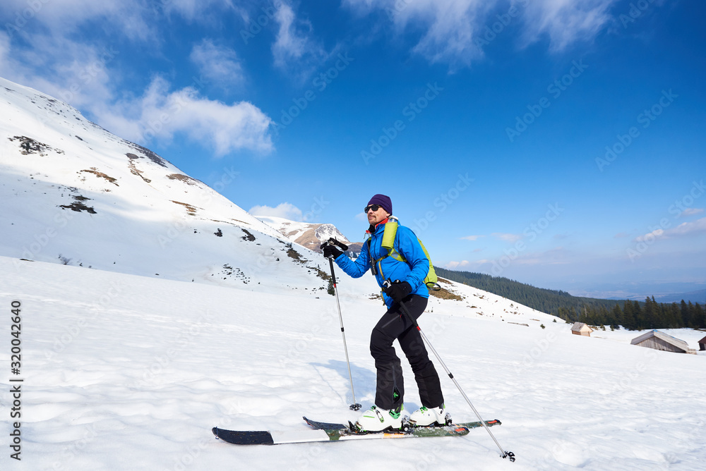 Handsome tourist in colorful clothing and sunglasses with backpack trekking on skis in deep snow on background of bright blue sky and beautiful mountain. Winter vacations, active lifestyle concept. Handsome tourist in colorful clothing and sunglasses with backpack trekking on skis in deep snow on background of bright blue sky and beautiful mountain. Winter vacations, active lifestyle concept.