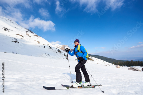 Handsome tourist in colorful clothing and sunglasses with backpack trekking on skis in deep snow on background of bright blue sky and beautiful mountain. Winter vacations, active lifestyle concept.