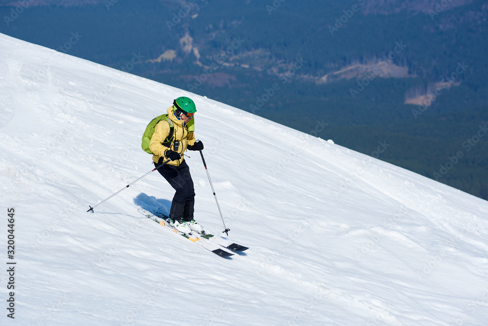 Professional skier in helmet with backpack riding skis fast down steep snowy mountain slope on background of deep white snow and green spruce trees. Winter holidays, sport and recreation concept.