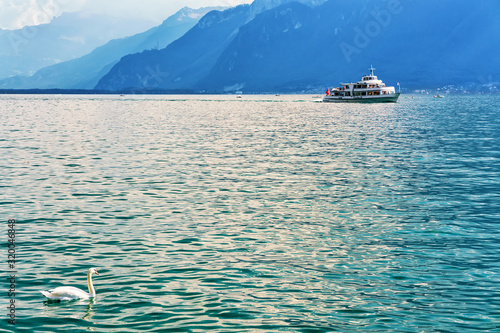 Swan and excursion ferry at Geneva Lake of Vevey, Swiss Riviera. Alps mountains and people on the background