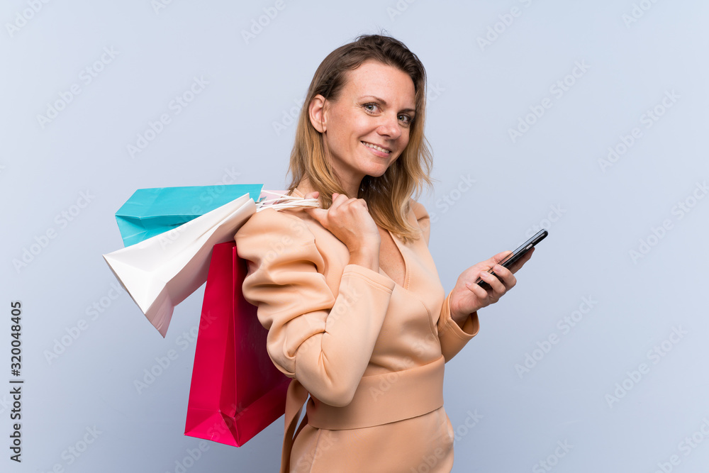 Naklejka premium Blonde woman over isolated blue background holding shopping bags and writing a message with her cell phone to a friend