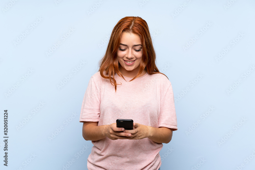 Teenager redhead girl over isolated blue background sending a message with the mobile Teenager redhead girl over isolated blue background sending a message with the mobile