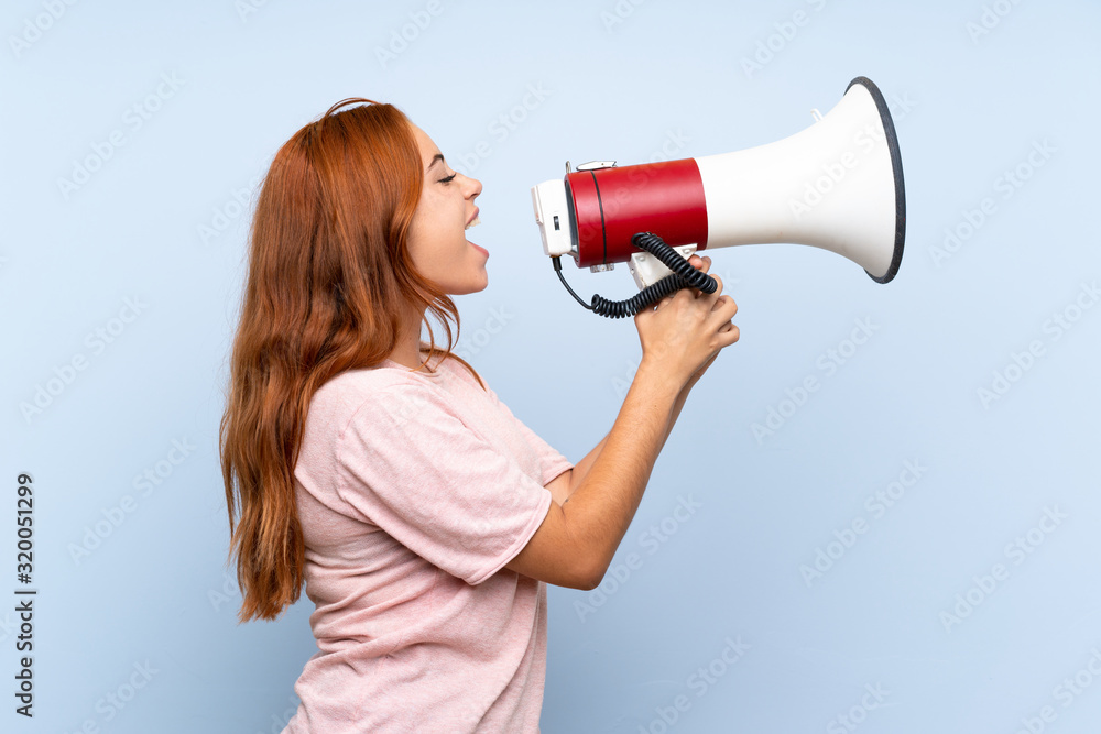 Teenager redhead girl over isolated blue background shouting through a megaphone Teenager redhead girl over isolated blue background shouting through a megaphone