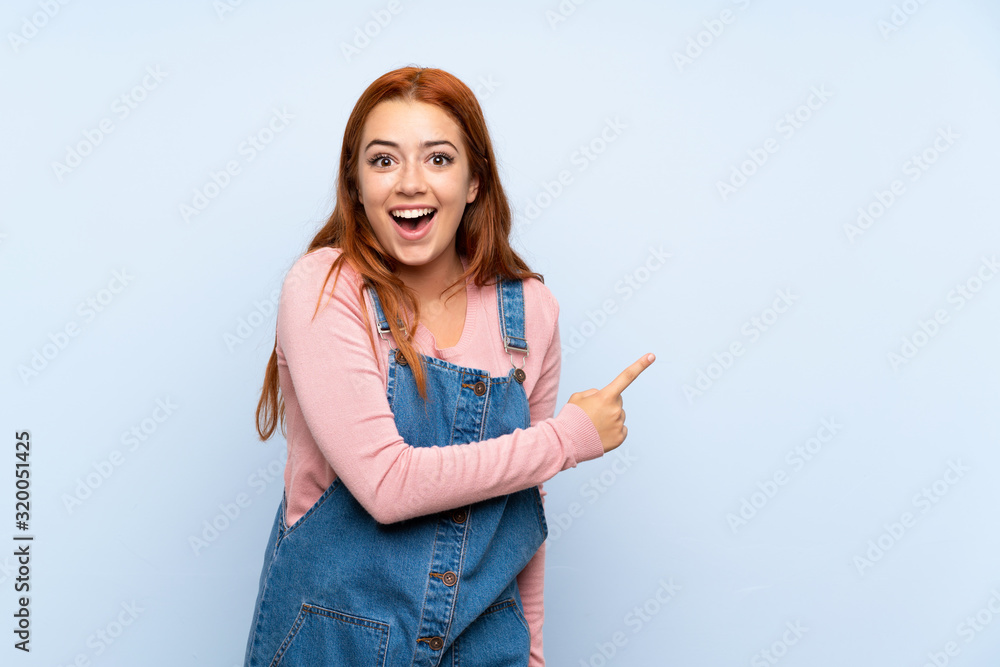 Teenager redhead girl with overalls over isolated blue background pointing finger to the side Teenager redhead girl with overalls over isolated blue background pointing finger to the side