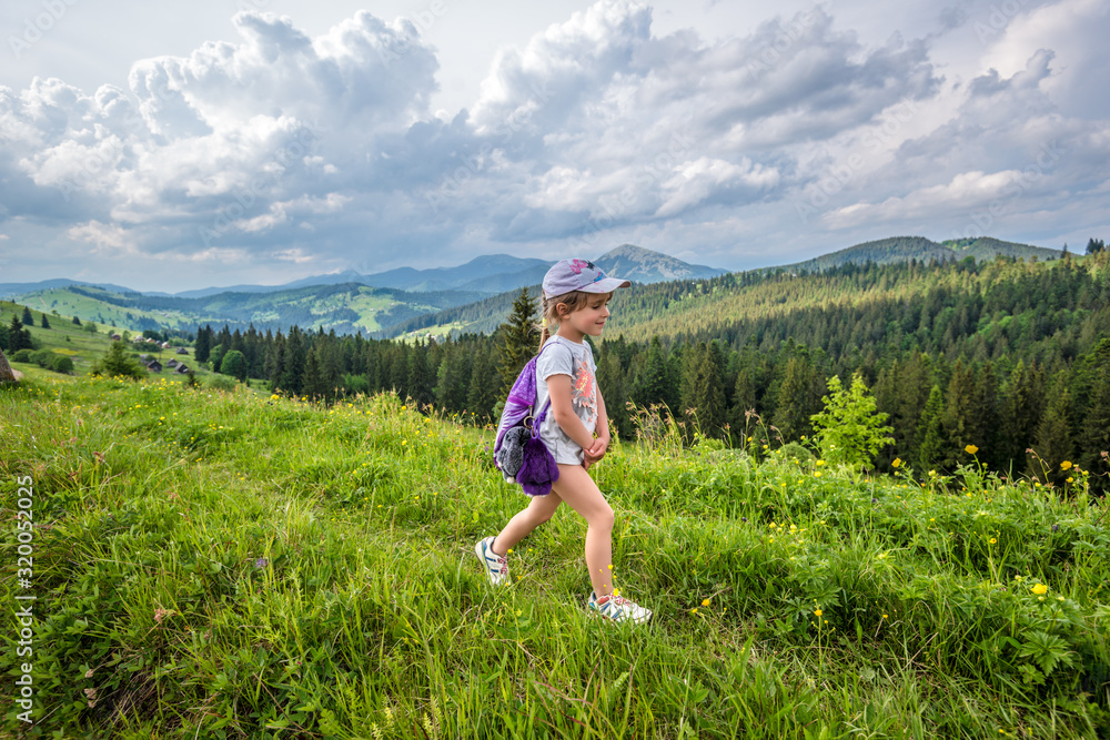 Side view little cute girl traveler lifts up a hill overgrown with green grass and meadow with flowers. Backdrop of picturesque hills and forest on a cloudy warm sunny summer day Side view little cute girl traveler lifts up a hill overgrown with green grass and meadow with flowers. Backdrop of picturesque hills and forest on a cloudy warm sunny summer day