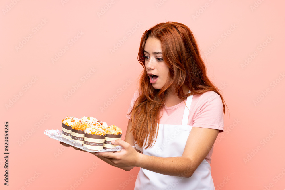 Teenager redhead girl holding lots of different mini cakes over isolated pink background Teenager redhead girl holding lots of different mini cakes over isolated pink background
