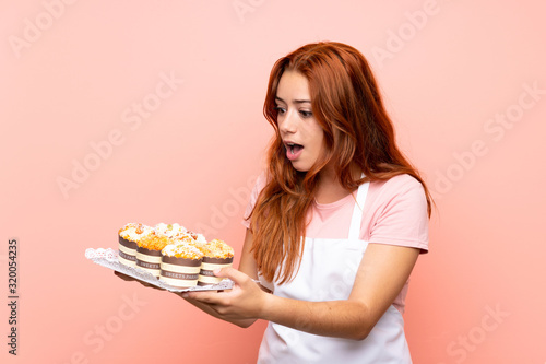 Teenager redhead girl holding lots of different mini cakes over isolated pink background