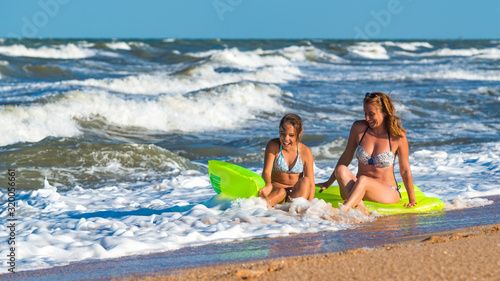 Positive young woman and her daughter bathe in the foamy stormy sea waves on an air mattress on a sunny summer day. Concept of enjoyment of relaxation and nature