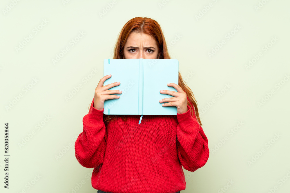 Teenager redhead girl with sweater over isolated green background holding and reading a book Teenager redhead girl with sweater over isolated green background holding and reading a book