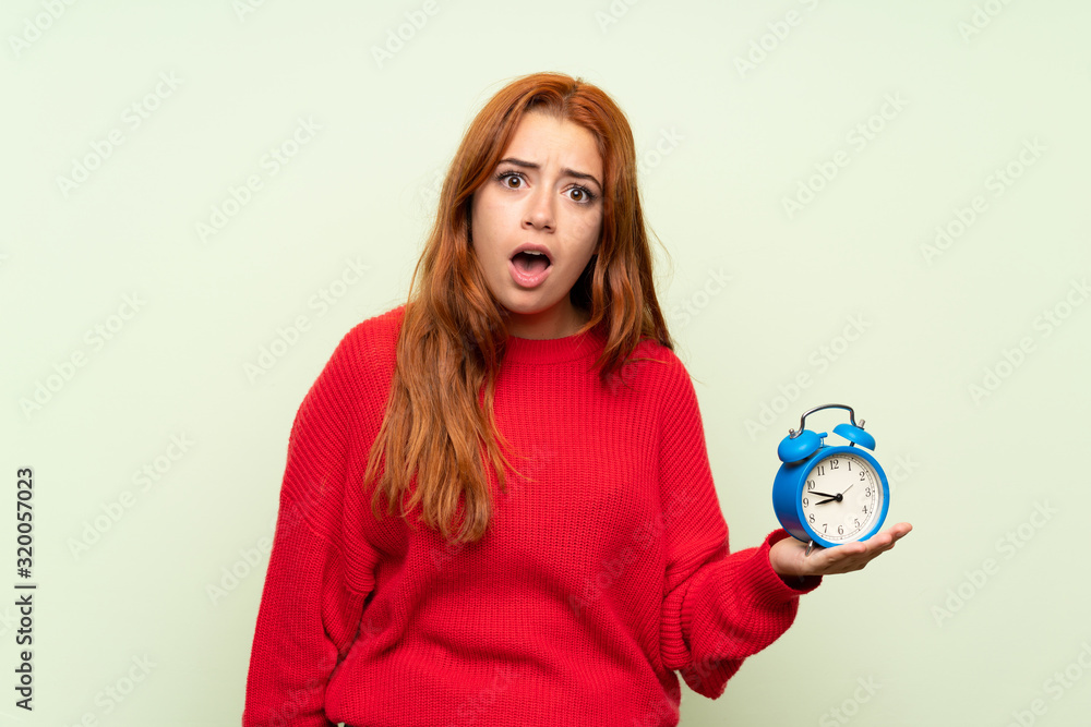 Teenager redhead girl with sweater over isolated green background holding vintage alarm clock Teenager redhead girl with sweater over isolated green background holding vintage alarm clock