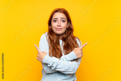 Teenager redhead girl over isolated yellow background pointing to the laterals having doubts
