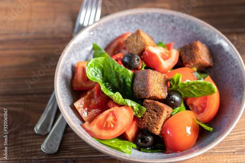 Italian salad of sliced tomatoes, basil leaves, olives, large pieces of croutons. In a deep plate, on a dark background.