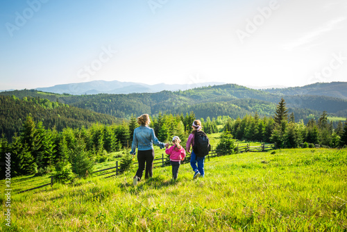 Rear view young mother and two daughters go down the hill overgrown with green grass. Gorgeous view of the forest growing on the mountains during trekking on a warm summer day