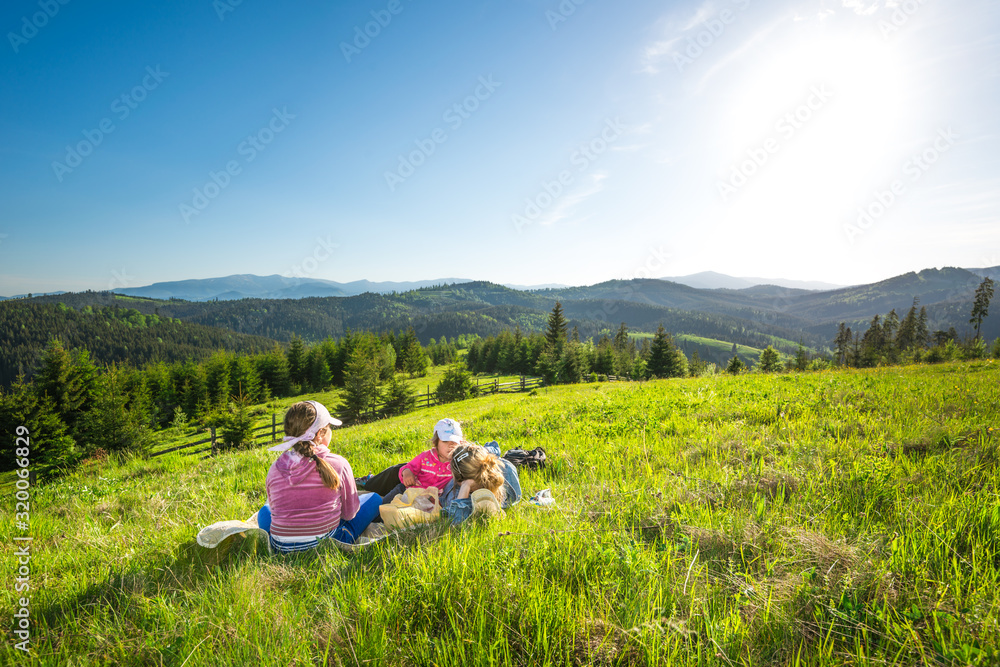 Young mother and little daughters lie on a slope overgrown with grass or admire the gorgeous view of a fox growing on the hills on a sunny warm summer day. Camping and trekking concepts. Copyspace Young mother and little daughters lie on a slope overgrown with grass or admire the gorgeous view of a fox growing on the hills on a sunny warm summer day. Camping and trekking concepts. Copyspace