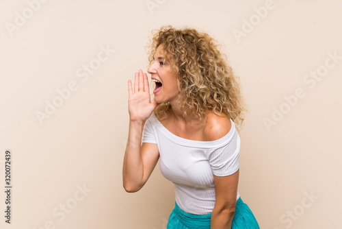 Young blonde woman with curly hair over isolated background shouting with mouth wide open to the lateral