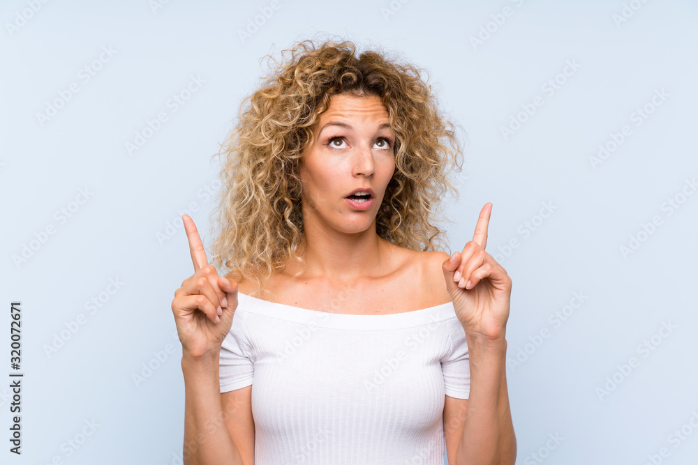 Young blonde woman with curly hair over isolated blue background pointing with the index finger a great idea Young blonde woman with curly hair over isolated blue background pointing with the index finger a great idea