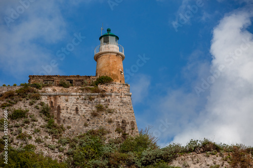 Lighthouse on the hill covered with green grass and plants on cloudy blue sky landscape background © Georgy
