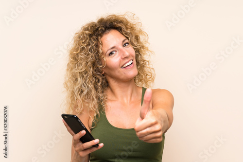 Young blonde woman with curly hair using mobile phone with thumbs up because something good has happened