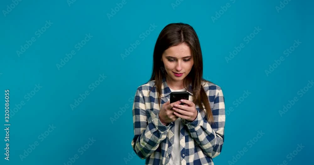 Front view of smiling girl in checkered shirt using smartphone isolated on blue background Front view of smiling girl in checkered shirt using smartphone isolated on blue background