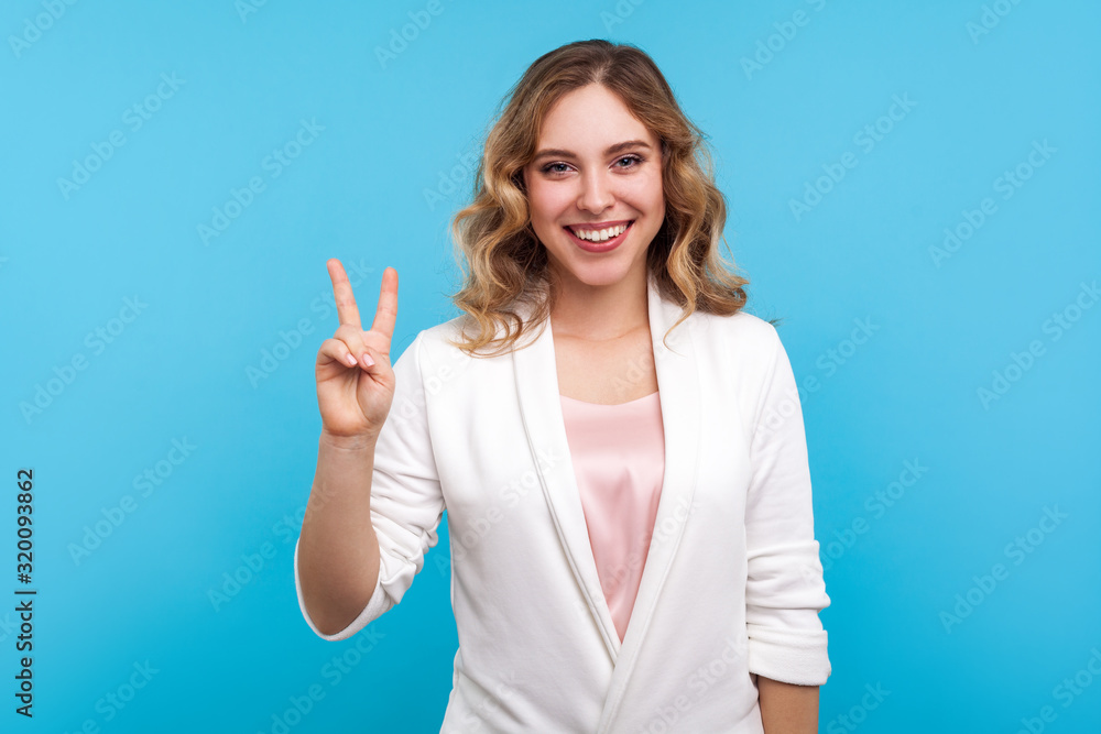 Victory! Portrait of smiling beautiful woman with wavy hair in white jacket showing v sign, peace gesture with two fingers, positive about winning and success. indoor studio shot, blue background Victory! Portrait of smiling beautiful woman with wavy hair in white jacket showing v sign, peace gesture with two fingers, positive about winning and success. indoor studio shot, blue background
