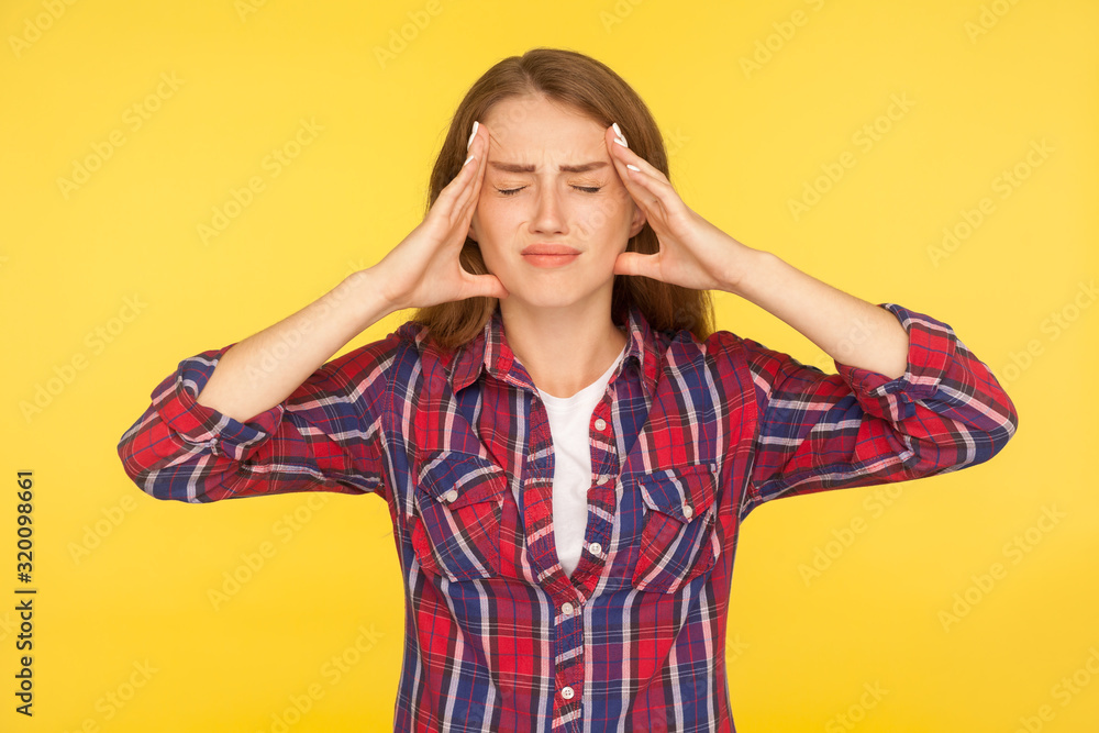 Headache. Portrait of stressed girl in checkered shirt clasping hands over sore head, grimacing from sudden pain having migraine, fever and flu symptoms. studio shot isolated on yellow background Headache. Portrait of stressed girl in checkered shirt clasping hands over sore head, grimacing from sudden pain having migraine, fever and flu symptoms. studio shot isolated on yellow background