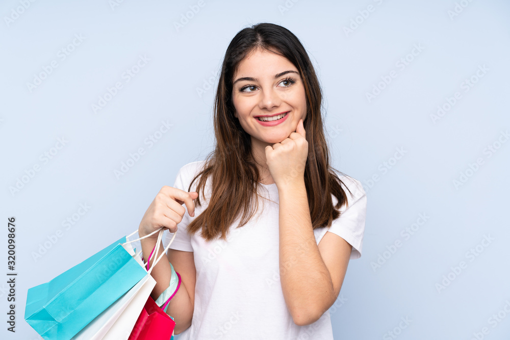 Young brunette woman over isolated blue background holding shopping bags and thinking Young brunette woman over isolated blue background holding shopping bags and thinking